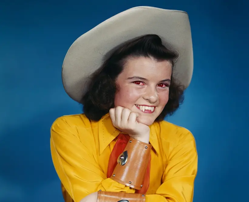 Colour photograph of a woman in cowboy attire posing with a gun in her hand. In the picture you can also see she has extended incisors and reddened eyes.