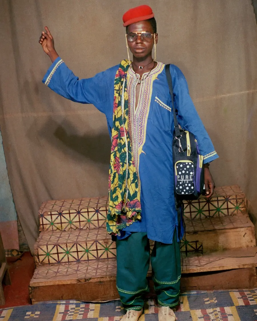 A young man stands in a makes-shift studio with one arm held aloft, pointing towards the ceiling. He wears a short, red felt cap, a long blue robe and green trousers with a backpack slung on one shoulder.