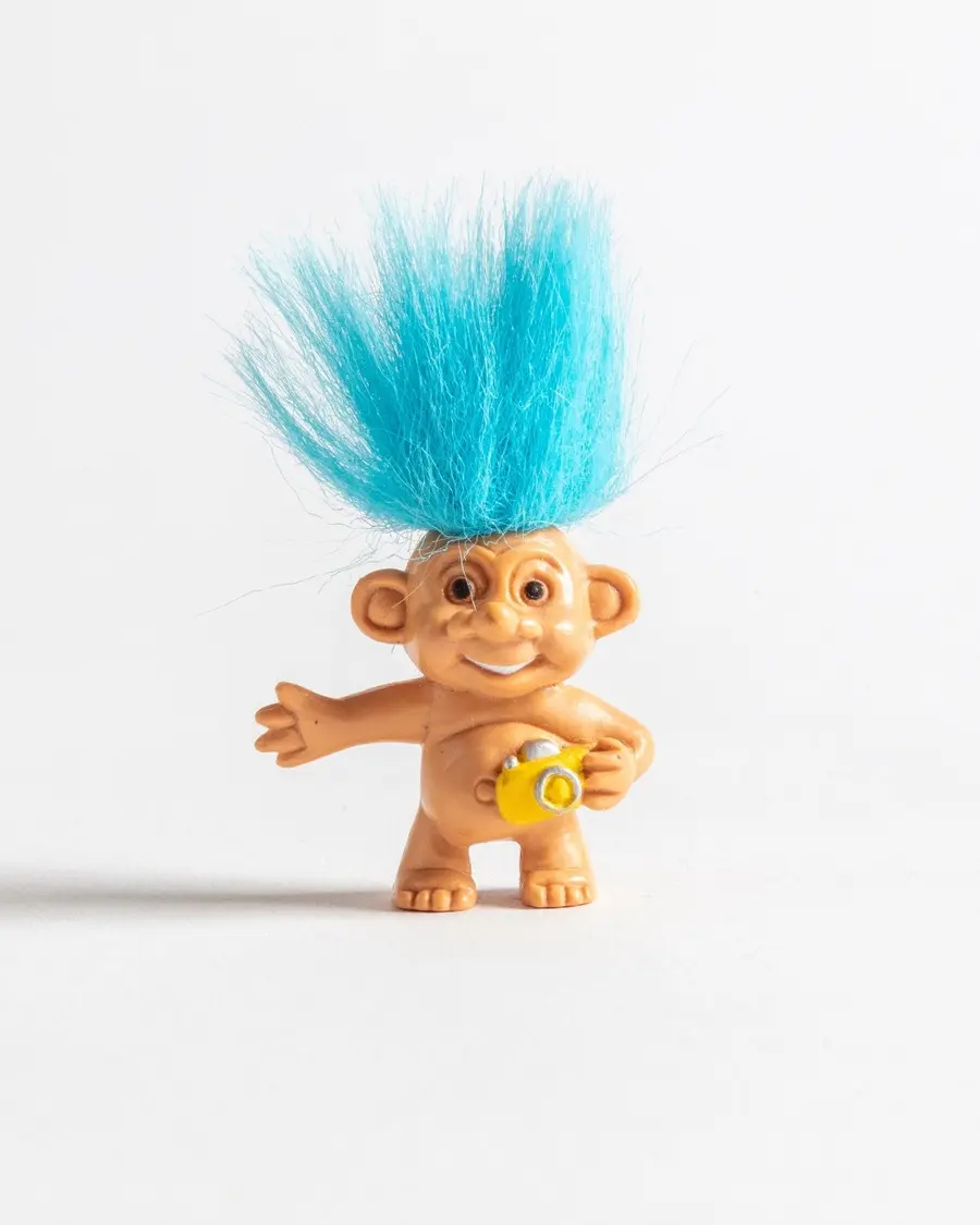 A plastic toy of a troll with blue hair and a huge smile holding a yellow camera