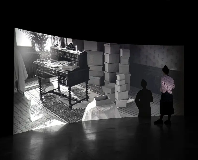 Installation photo of a screen showing a room full of boxes. A person can be seen watching this screen.