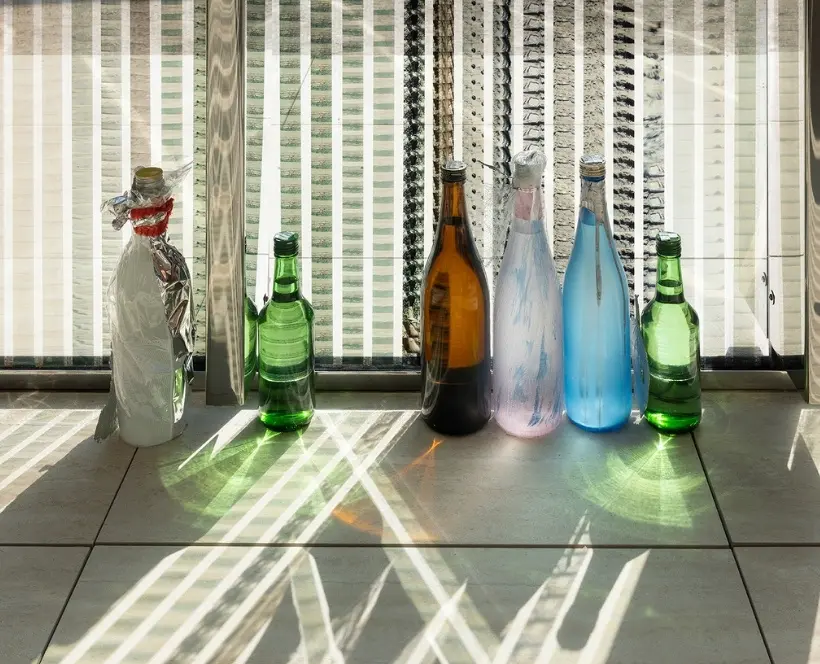 Installation image of Lotus L Kang’s work in situ at the Giardini della Biennale, Venice. In the photo, there are multiple glass bottles arranged next to a window.
