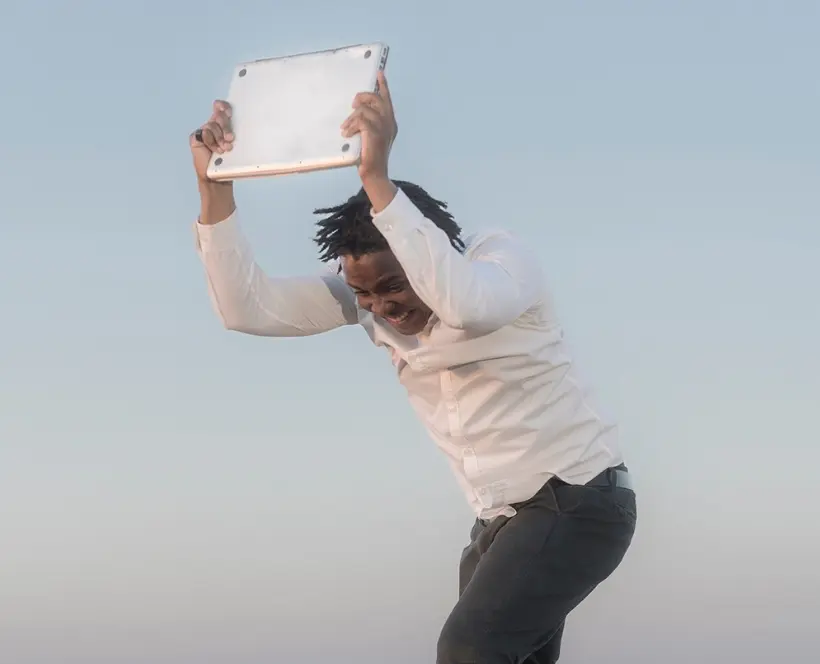 Colour film still of a man holding an ipad in his hands with the aim of hitting it against the rocky ground that he stands on.