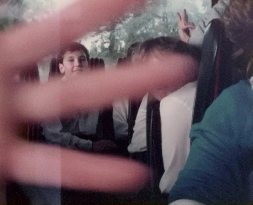 A blurry, close-up hand with fingers spread dominates the foreground, obscuring most of the view of several people seated in a vehicle. One person behind the hand smiles, and another's hand makes a peace sign in the upper right.