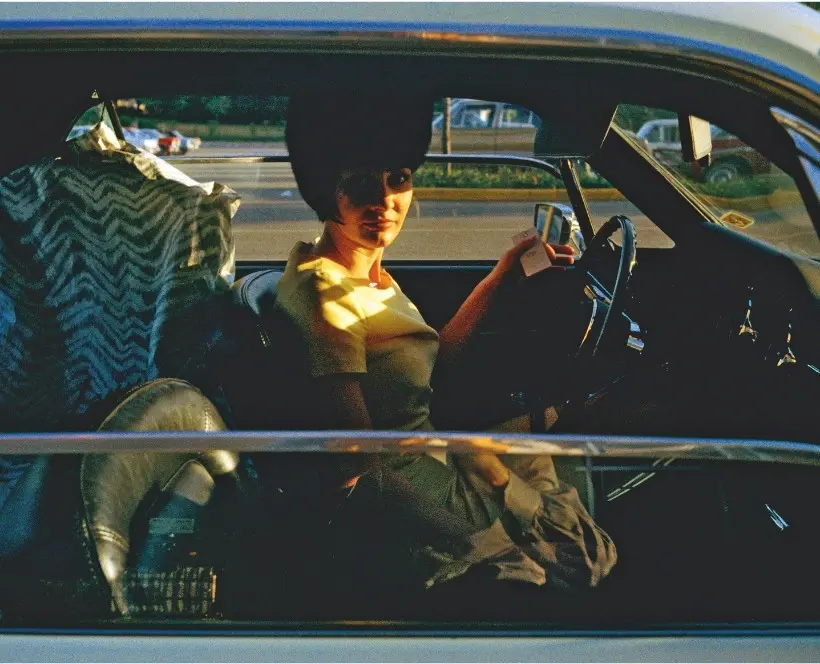 Colour photograph of a woman sat in the driver’s seat of a car with clothes hung up behind her.