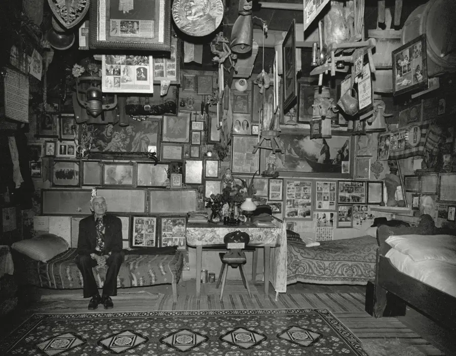 Black and white image of an elderly Polish man sat inside a room covered in ephemera both hung on the walls and from the ceiling. 