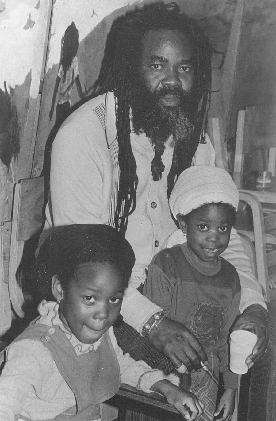 A black and white photograph of a man in a white shirt, sat behind his two young children, holding one of them in his lap.