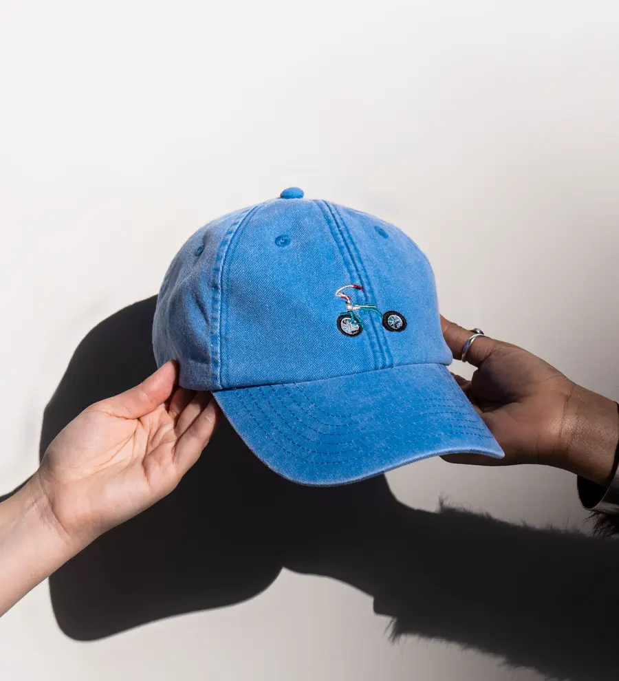 photograph of a blue cap with a tricycle embroidered on the front. Two hands are holding the cap up