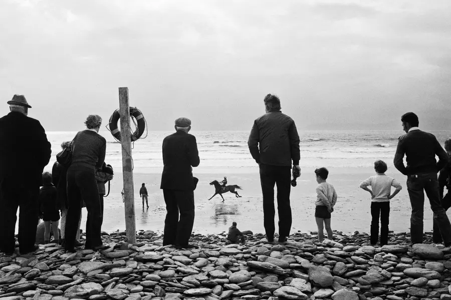 Black and white photograph showing a group of people standing on a rocky shore watching a person riding a horse.