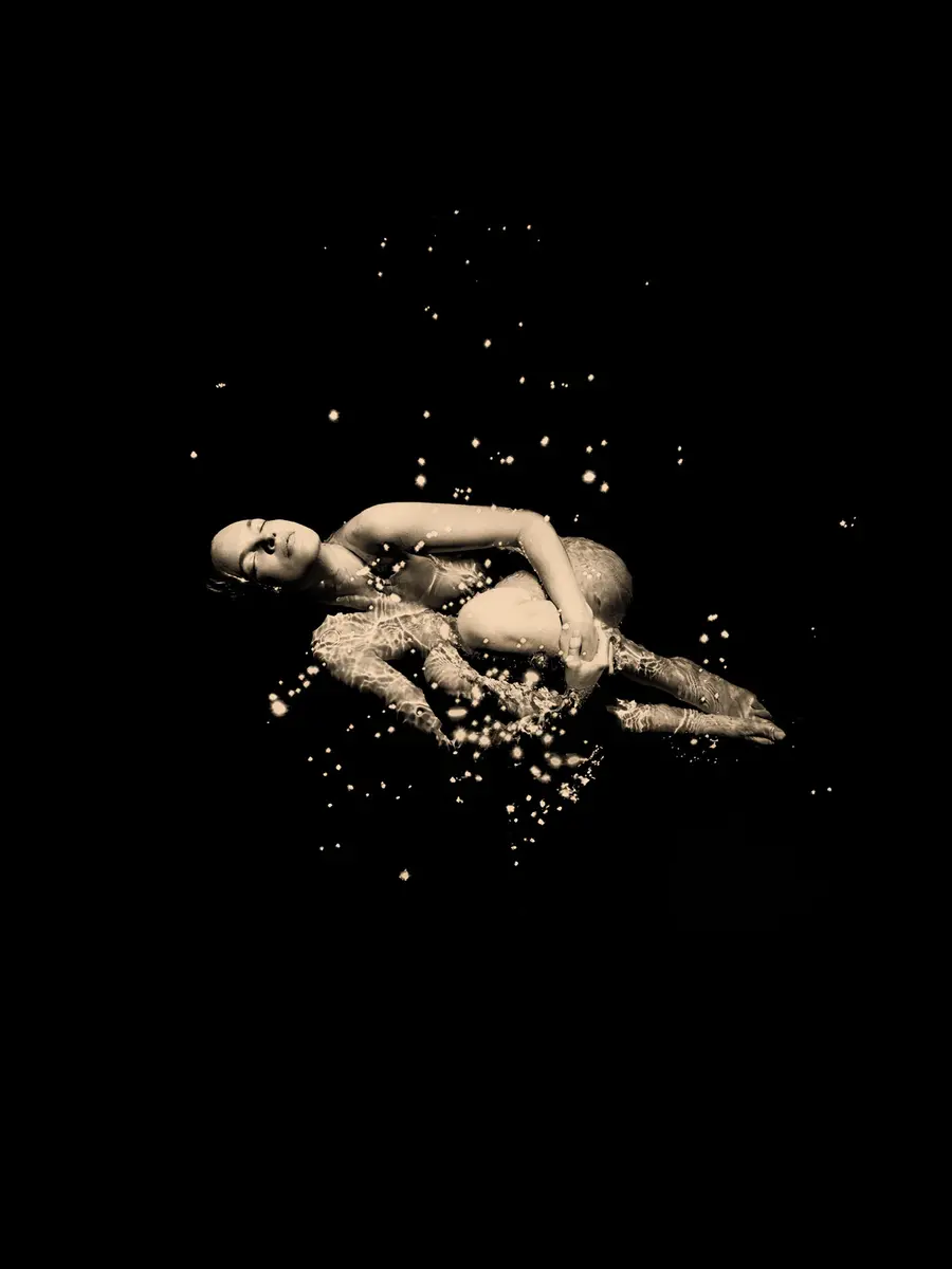 A black and white image of a woman curled up into a fetal position floating in a pool of dark water with a flurry of cherry blossom floating about her.