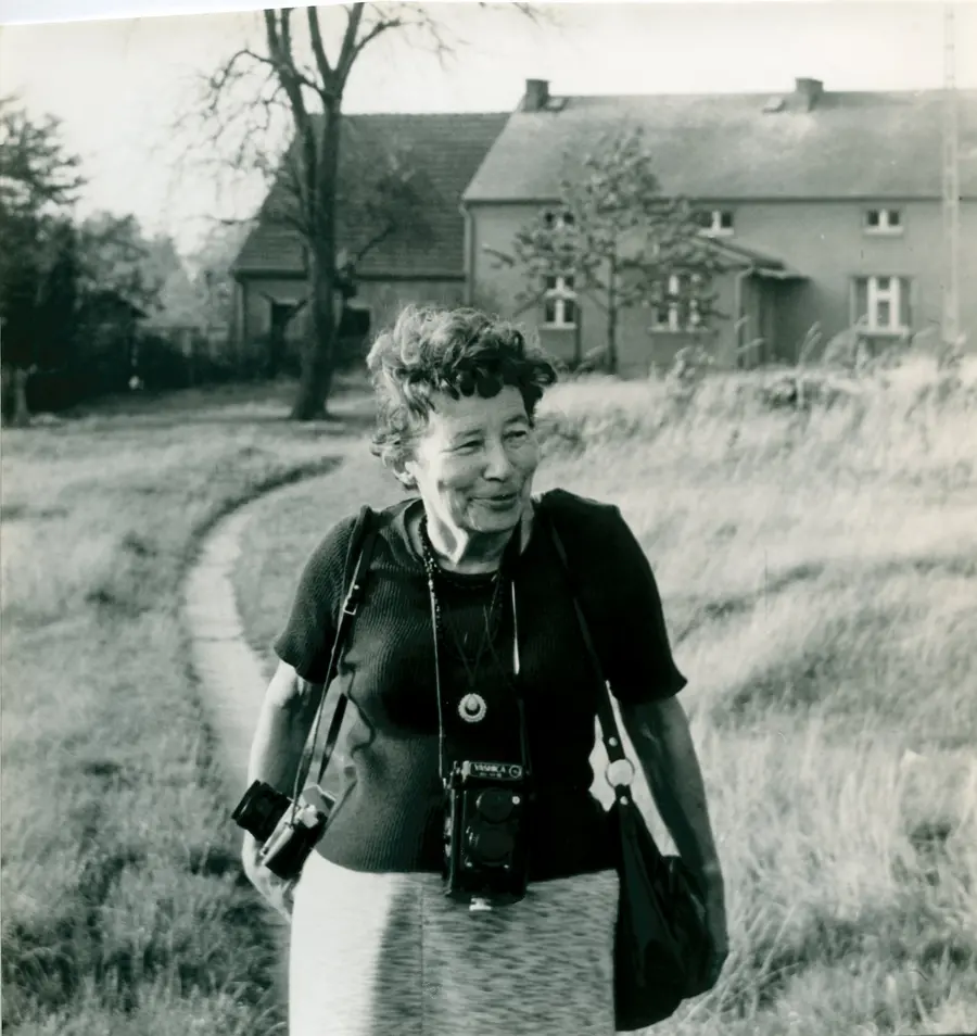 Black and white photograph of Zofia Rydet walking down a path leading from a traditional country cottage. She wears a dark blouse and has a camera on a strap slung around her shoulder.