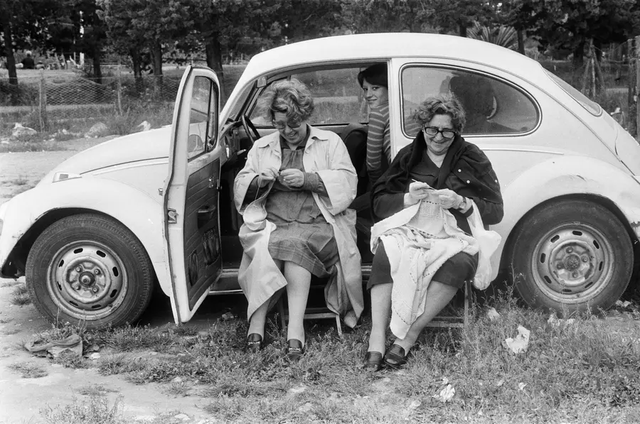 two older women sit on folding chair in front of a car.