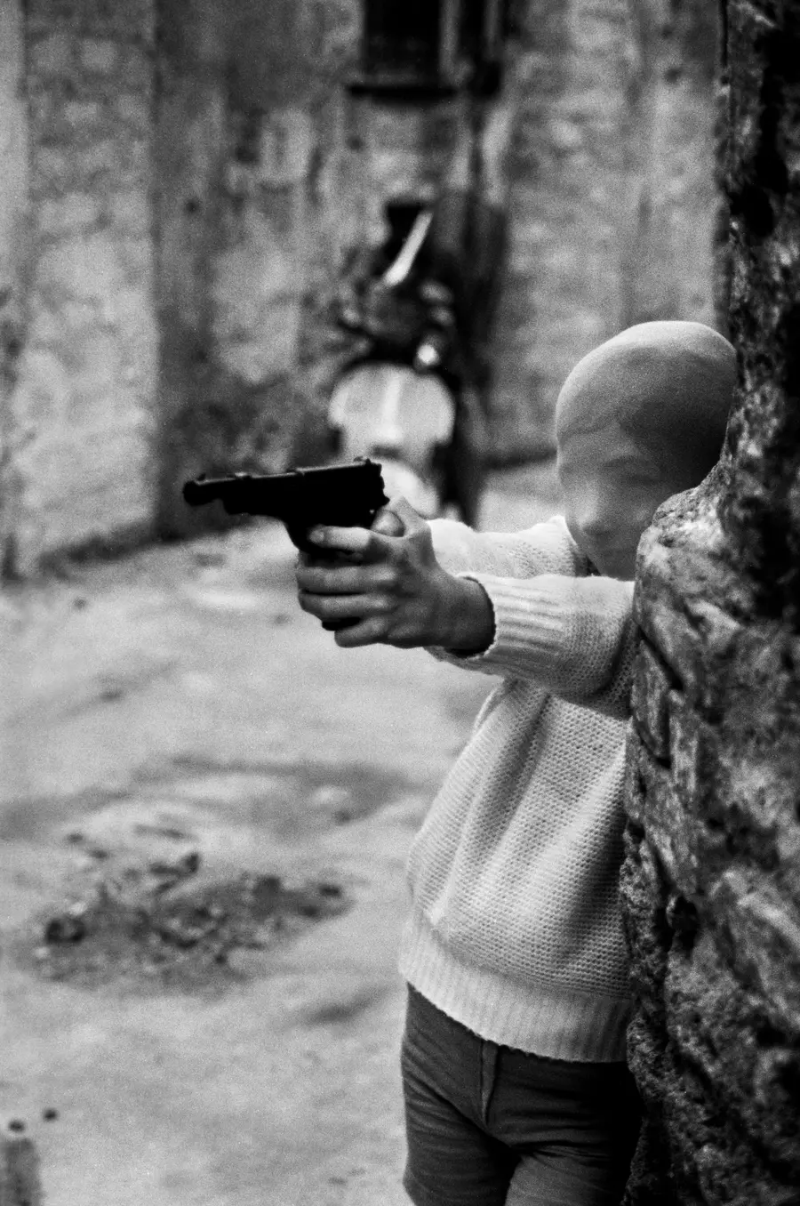 Black and white photograph of a young boy with a thin balaclava-like disguise over his head. He is aiming a handgun at something. 
