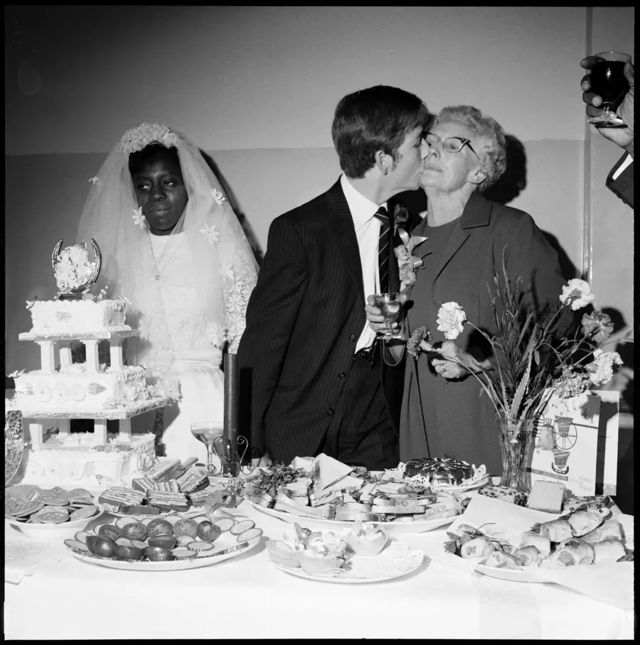 A young couple on their wedding day. The woman stares out over the cake with her wedding veil pulled back as her groom leans away to kiss his mother on the cheek.