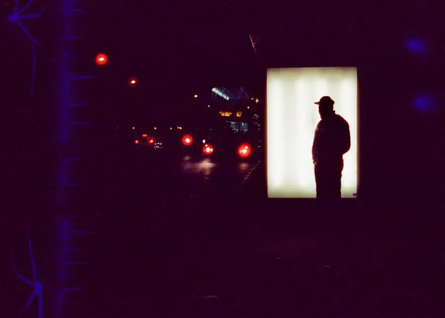 Colour photograph taken at night with a figure stood silhouetted against the bright light of a bus stop stand