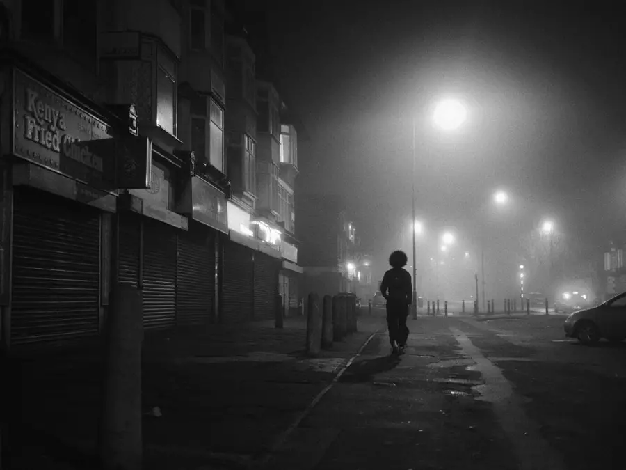 Black and white photograph of a night street scene with a figure walking away from the camera on the street on a foggy night with street lights along the street