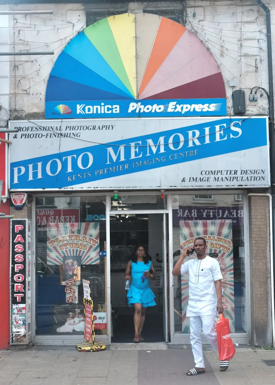 Colour photograph of a man and woman stood outside a shop with a rainbow effect semi-circular sign above the shop which has a sign Photo Memories as the name of the shop