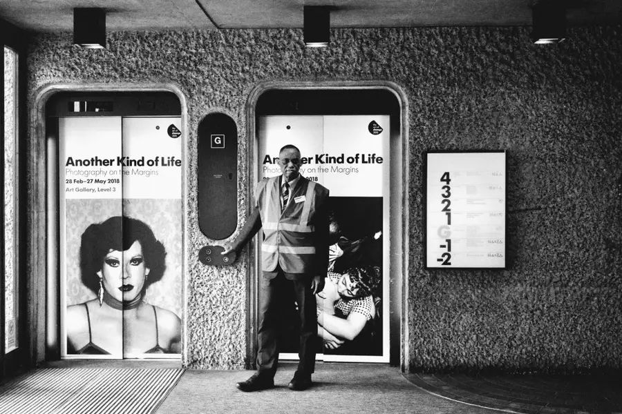 Black and white photograph of a man wearing a suit with a high visibility vest and name badge, stood next to two lifts. The doors of the lifts are displaying adverts for an exhibition at the Barbican 