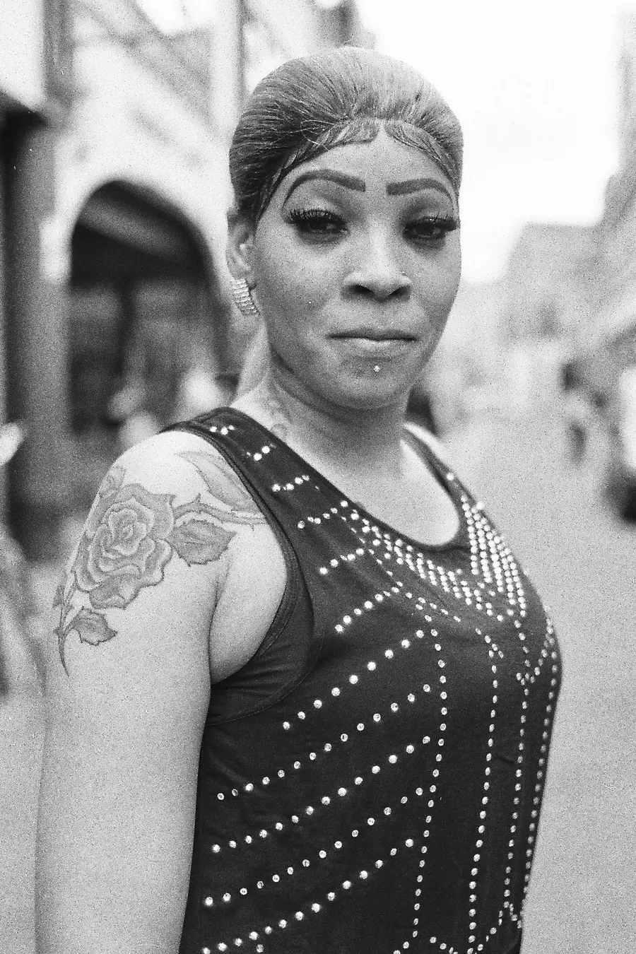 Black and white photograph of a portrait of a woman stood looking at the camera, wearing a sleeveless top with a tattoo in the design of a rose on the top of her right arm
