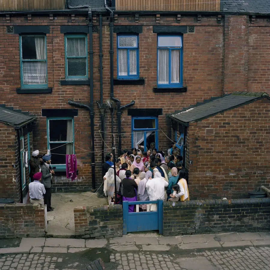 Colour image of a gathering of women huddled with each other outside the back of some terrace houses. A smaller group of men stand waiting with cameras to the side to capture some group photographs.