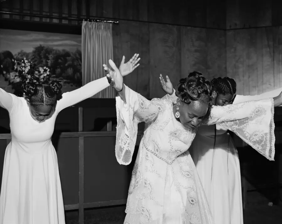 Black and white photograph of three women, wearing white dresses. They have their heads bent down with their arms outstretched.