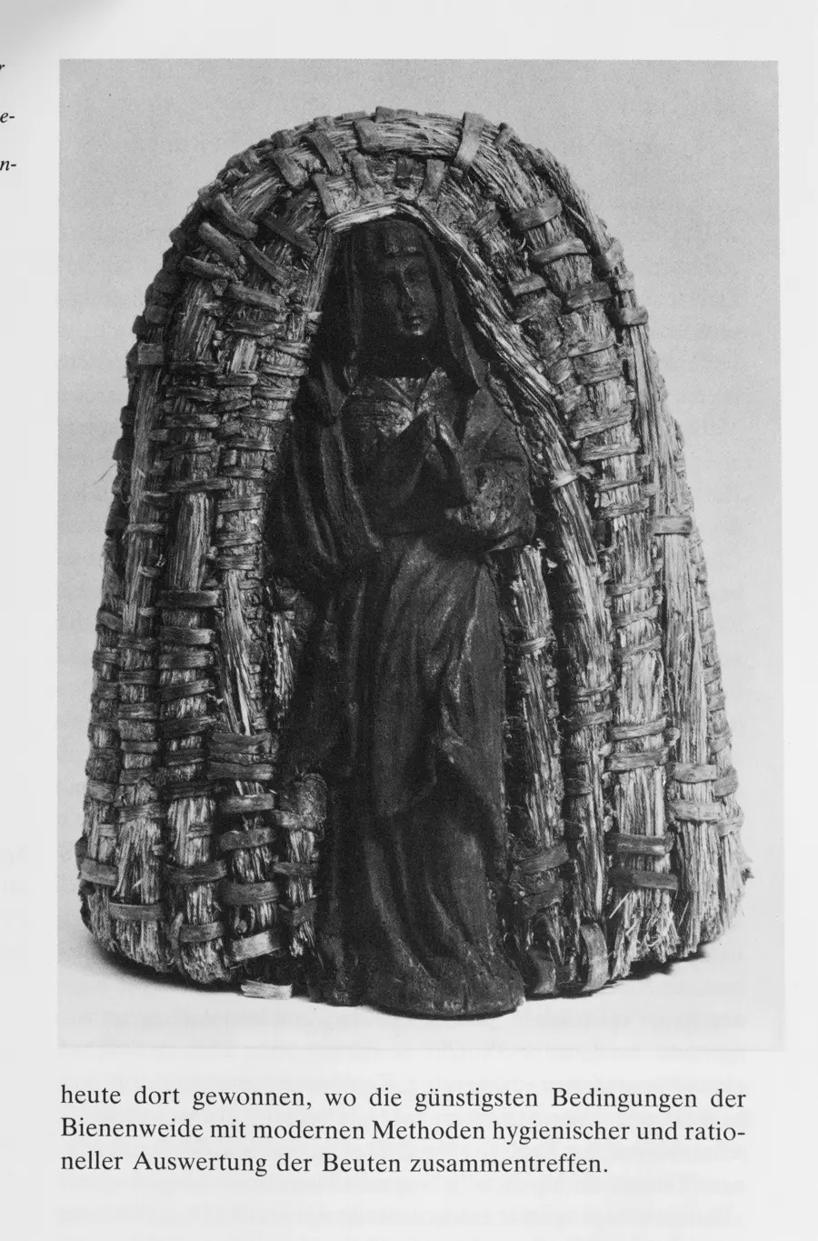 A black and white scan of a page of a book showing a beehive made of woven rushes with a the figure of a Madonna in front of it 