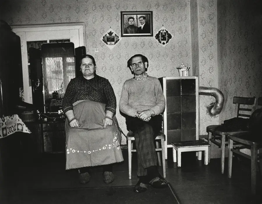 A black and white image of an elderly couple sat beneath a portrait of themselves as young adults at their wedding. 