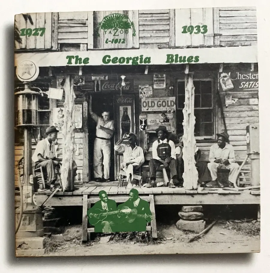 The cover of a 1968 compilation album titled ‘The Georgia Blues 1927-1933’ featuring a black and white photograph by Dorothea Lange of a group of men sitting on the porch of a general store, with the album title and an additional image superimposed on spaces in the image. 