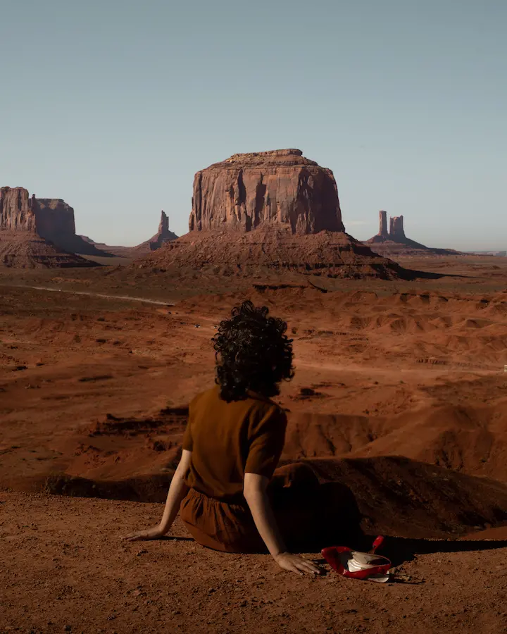 A woman sits with back to camera wearing a brown dress in a predominantly brown desert landscape with a pale blue sky. 