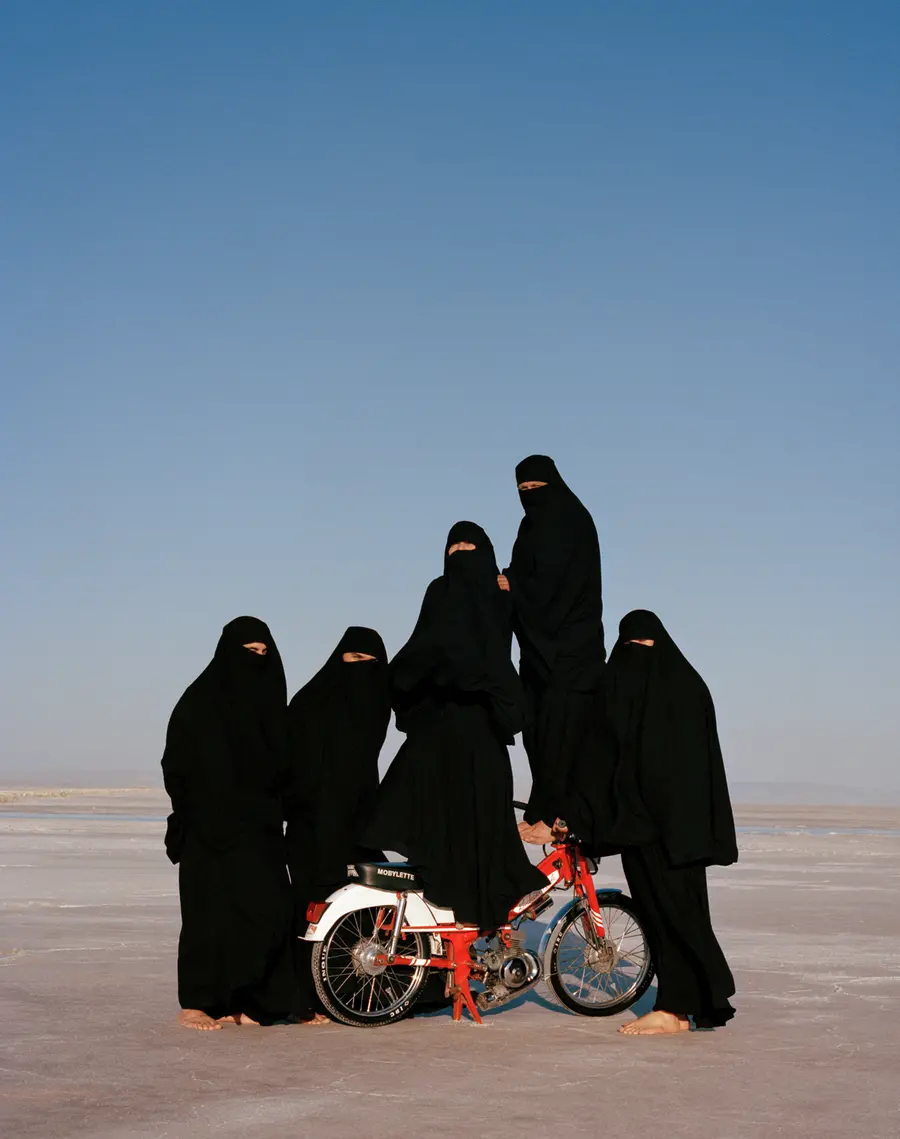 Five figures dressed in black hijabs stand in the desert with two balanced on a motor scooter.
