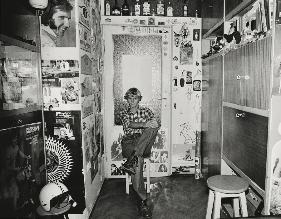 Black and white photograph of a young man  sat on a stool in a room full of signs and stickers.