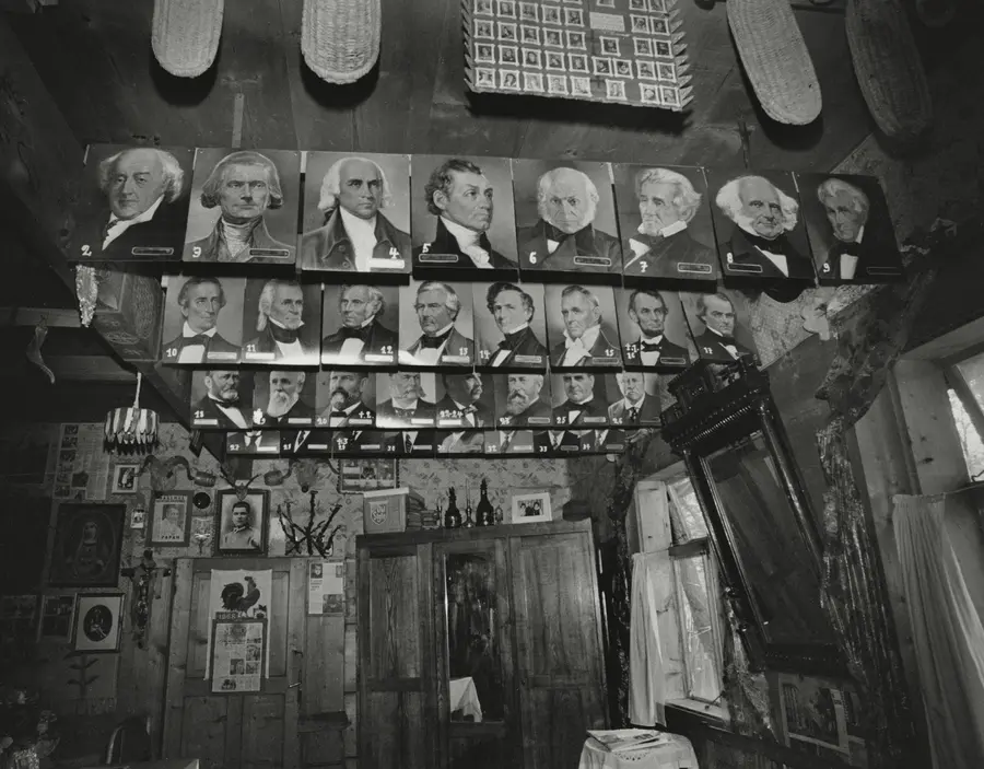 Black and white photograph of a room  featuring several photographs of men in formal suits  hanging in rows. On the walls, more photographs and  newspaper clippings can be seen along with other objecs  placed on top of a wardrobe