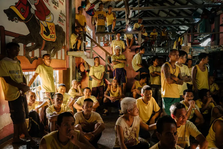 A group of over 100 men, crowded together in a large warehouse space and most wearing yellow t-shirts with 'Manilla City Jail Detainee' on the front, look to camera either standing or seated.