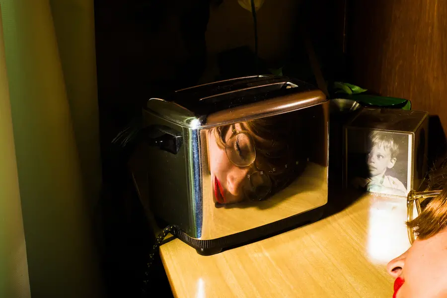 Reflection in the side of a toaster of a woman's head resting sideways on a countertop.