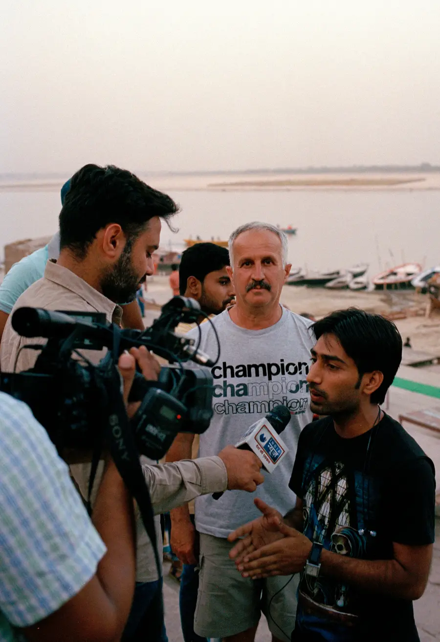 A man stands next to a person being interviewed on video camera, looking directly at the photographer's camera.