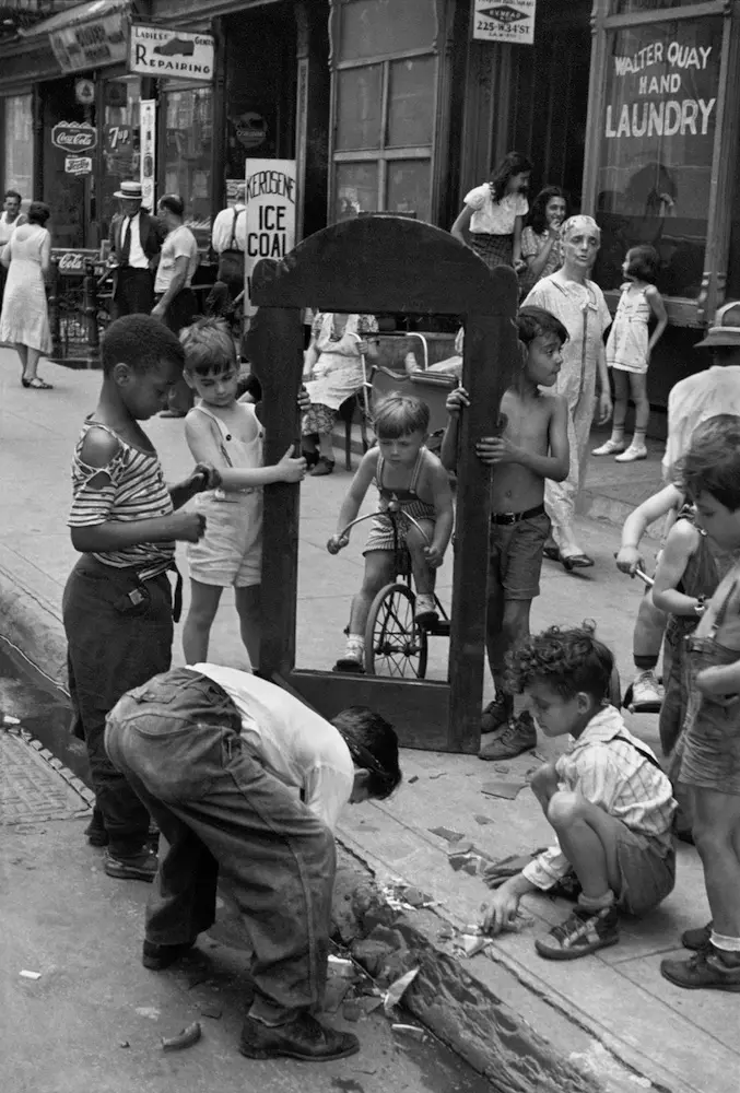 Nine children stand and squat around the large empty frame of a broken mirror on a busy city pavement.