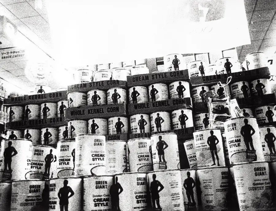 Black and white photograph of stacks of canned goods, with a silhouetted figure on the label