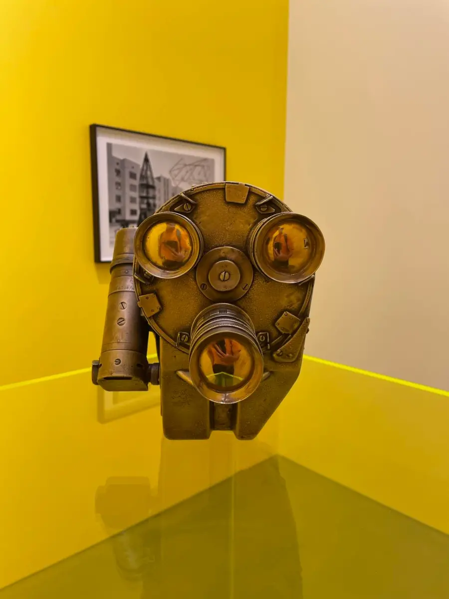Image of old camera on yellow podium, the camera has three lenses, resembling a face or gas mask