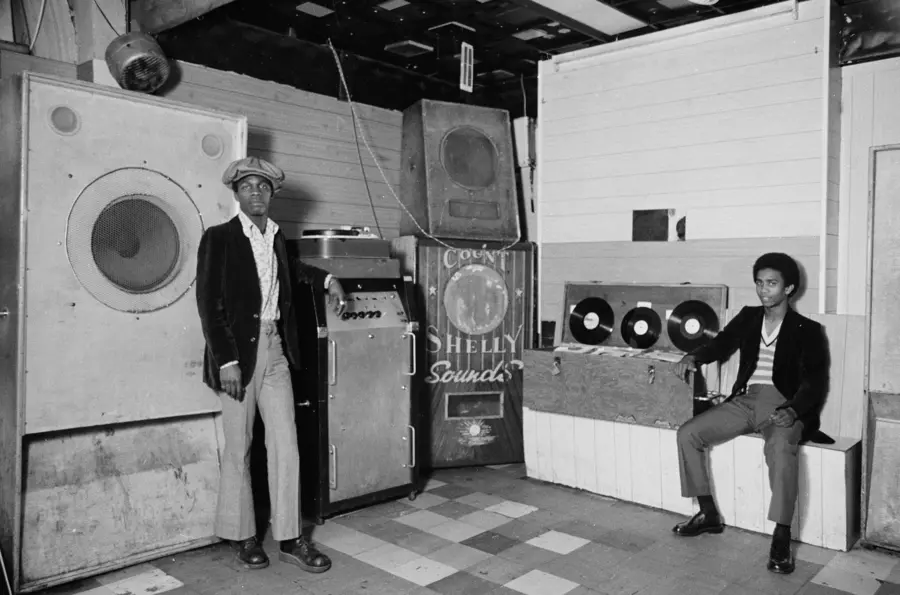 Two young black men sit in a basement club surrounded by sections of a sound system.