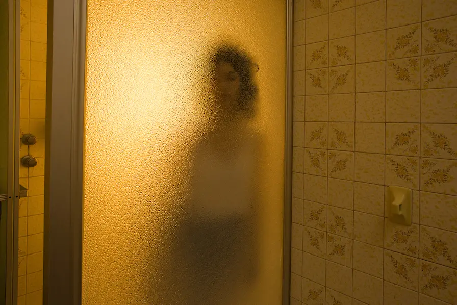 A clothed figure stands behind a mottled translucent shower door in a predominantly yellow photograph.