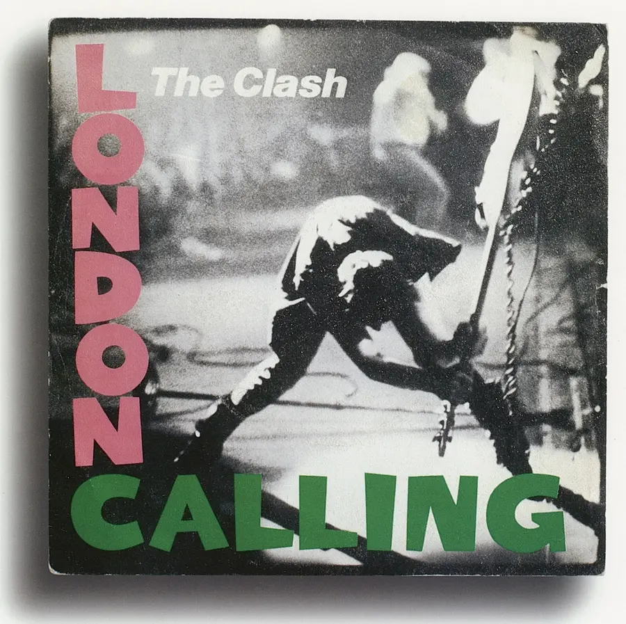 The album cover for ‘London Calling’ by The Clash combines a grainy black and white photograph of bassist Paul Simonon leaning down to smash his guitar to the stage floor with pink and green bold lettering. 