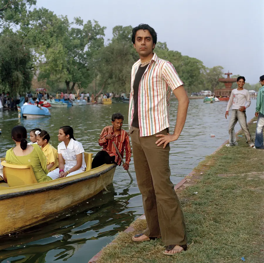 Figure standing on a riverbank looking to camera as a small pleasure boat floats alongside.