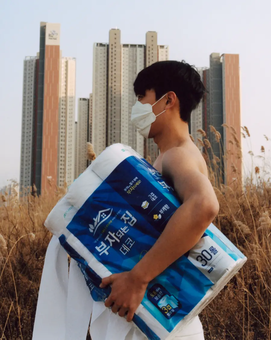 A man wearing a facemask but no shirt walks through a field with a nine-roll package of toilet paper under his arm.