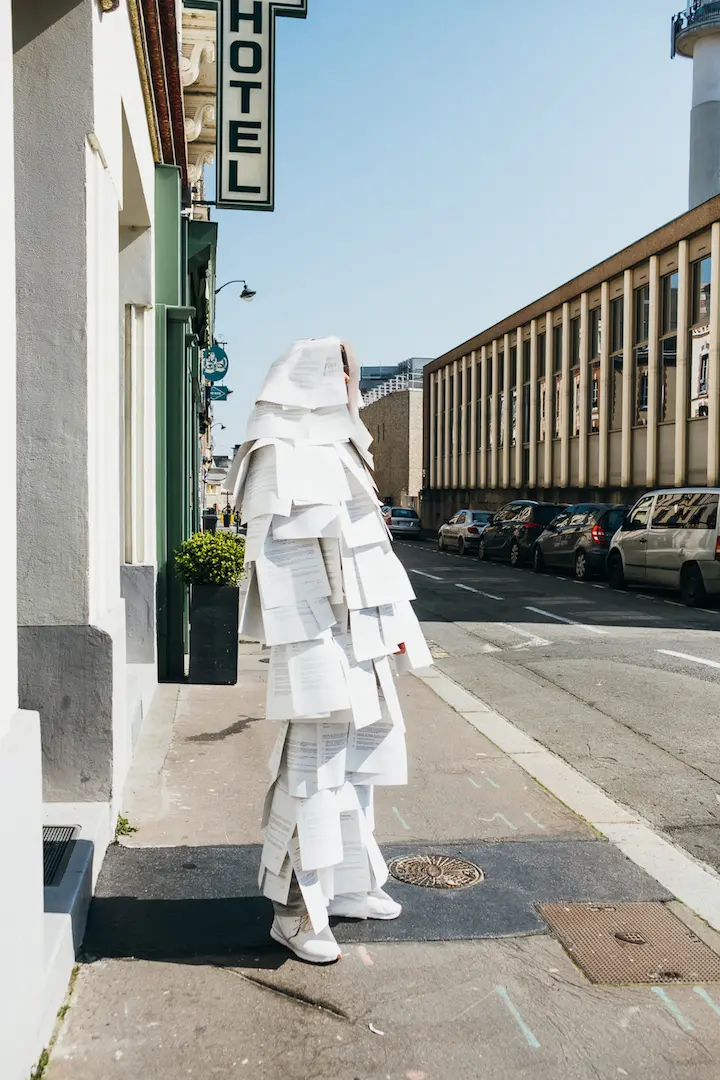 A figure stands outside a building on a street, they are covered in white sheets of paper with black text.