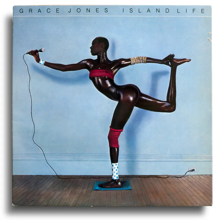 Cover of the album cover for Grace Jones 'Island Life' featuring a full length side portrait of a tall, short-haired black woman facing left. She balances touching her right leg, folded up and behind her, with her outstretched right arm. Her right arm is stretched in front holding a microphone. She is nude but for a thin red strip of fabric across her breasts and strips of colourful fabric on her wrists, elbow and knee. The wall behind her is pale blue. 