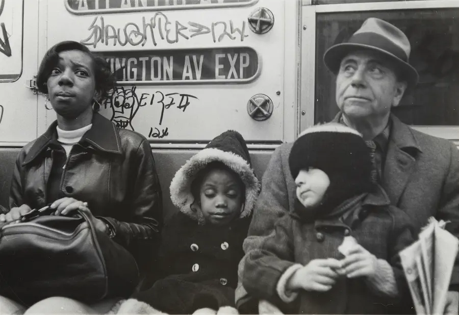 A man and a woman sit side by side facing the camera on a subway carriage, with two small children - one on the man's lap, the other between them.