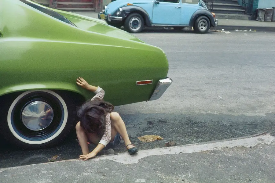 A girl squats beside the rear tyre of an avocado green car, body and face twisted away from the camera.