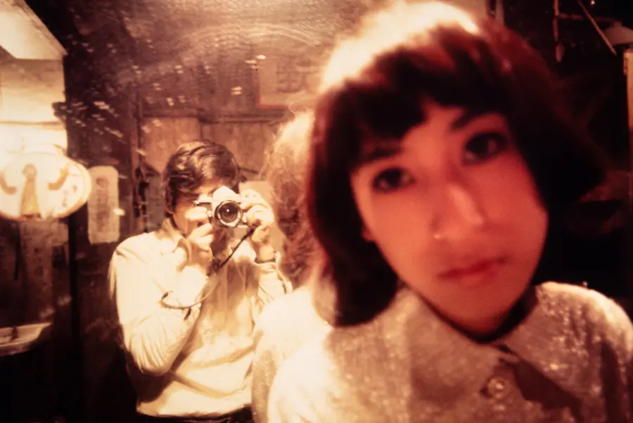 Colour photograph taken in a mirror of a blurred face with the photographer reflected in the background