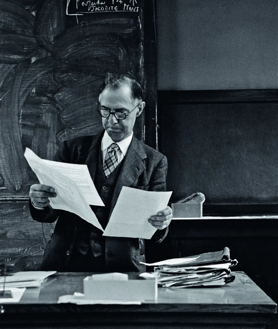 Black and white photograph of a man holding sheets of paper in his hand over a table.