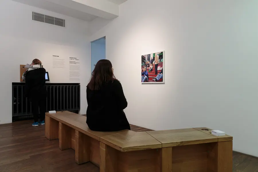Woman sitting on a bench looking at a framed photograph of a woman sitting on stairs, while another person in the room posts a postcard in a box.