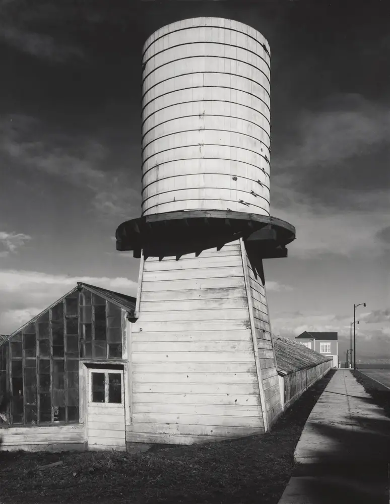 Black and white photograph of a wood-paneled water tower attached to a greenhouse in a landscape.
